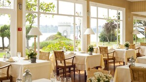 A dining area with tables set for a meal, large windows with a view of greenery and a distant structure, and wicker chairs. - Le Grand Hotel des Bains & Spa - Bretagne (Locquirec)