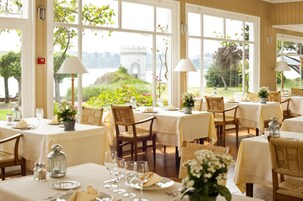 Un comedor con mesas dispuestas para una comida, ventanas amplias con vistas a zonas verdes y una estructura distante, y sillas de mimbre.