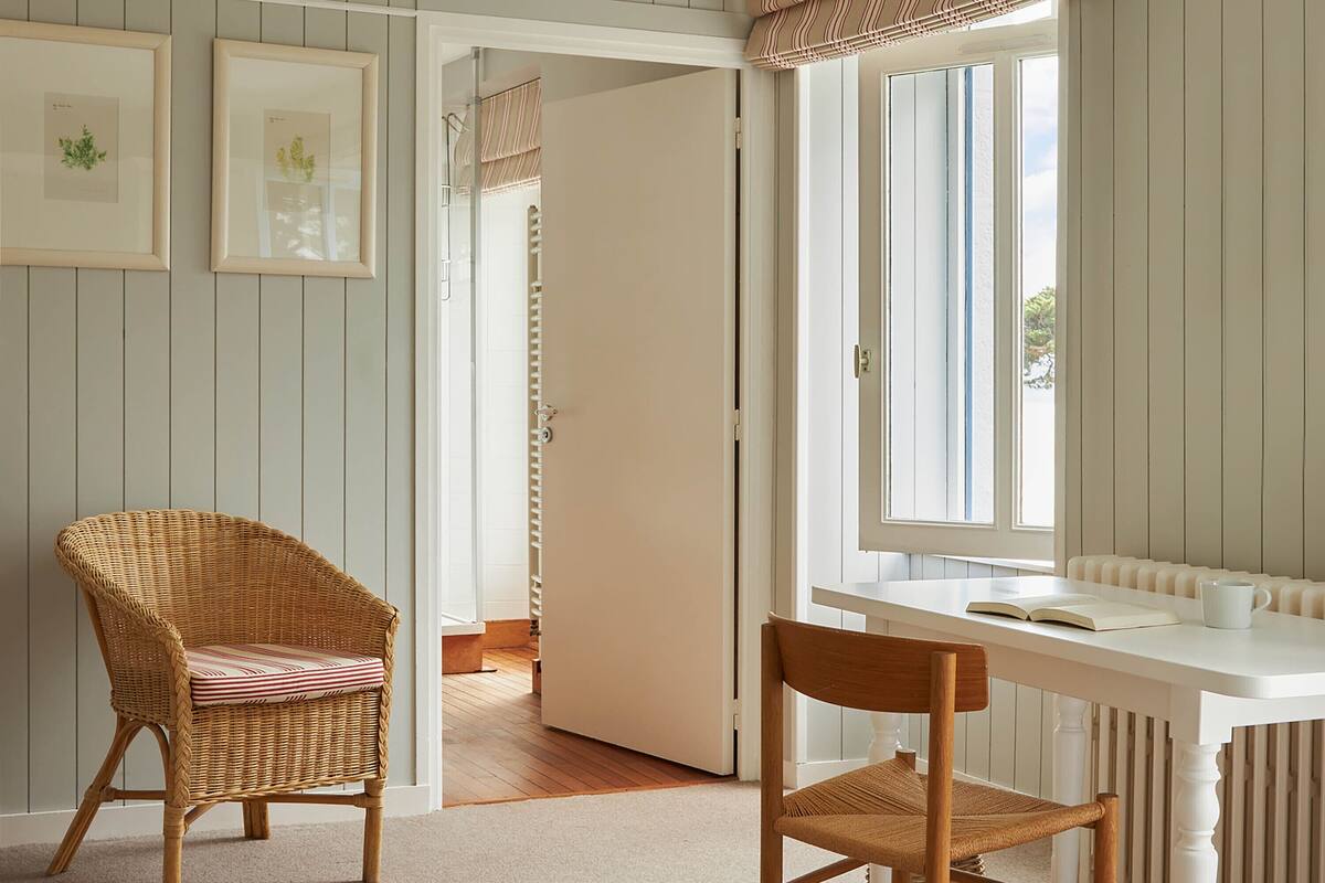A room with a wicker chair, a white table, and a window with striped curtains.