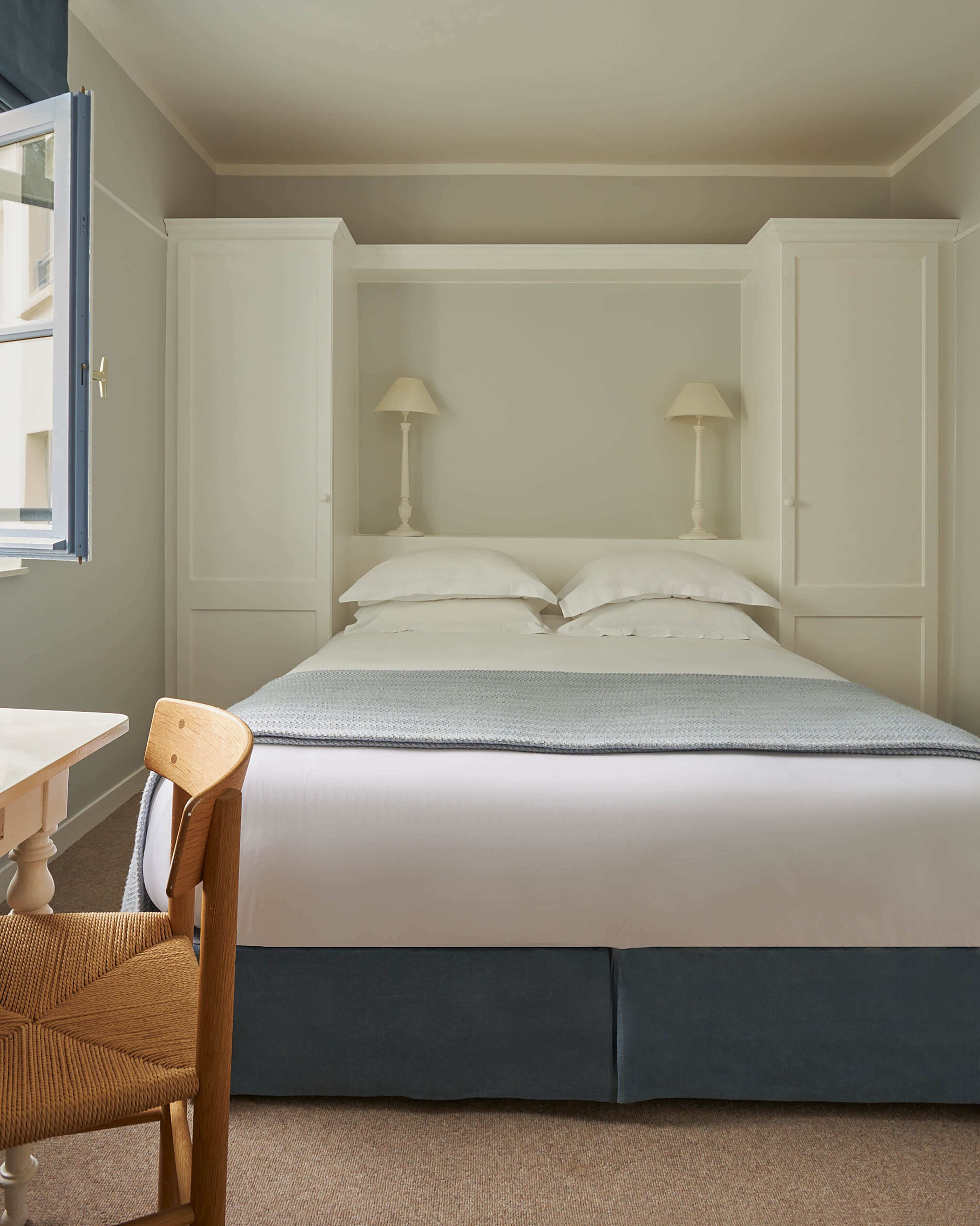 A neatly made bed with white linens, a grey blanket, and two bedside lamps. There is a wooden chair with a woven seat and a small table with a white surface. The room has a beige carpet and white walls.
