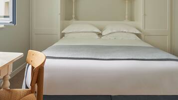 A neatly made bed with white linens, a grey blanket, and two bedside lamps. There is a wooden chair with a woven seat and a small table with a white surface. The room has a beige carpet and white walls.
