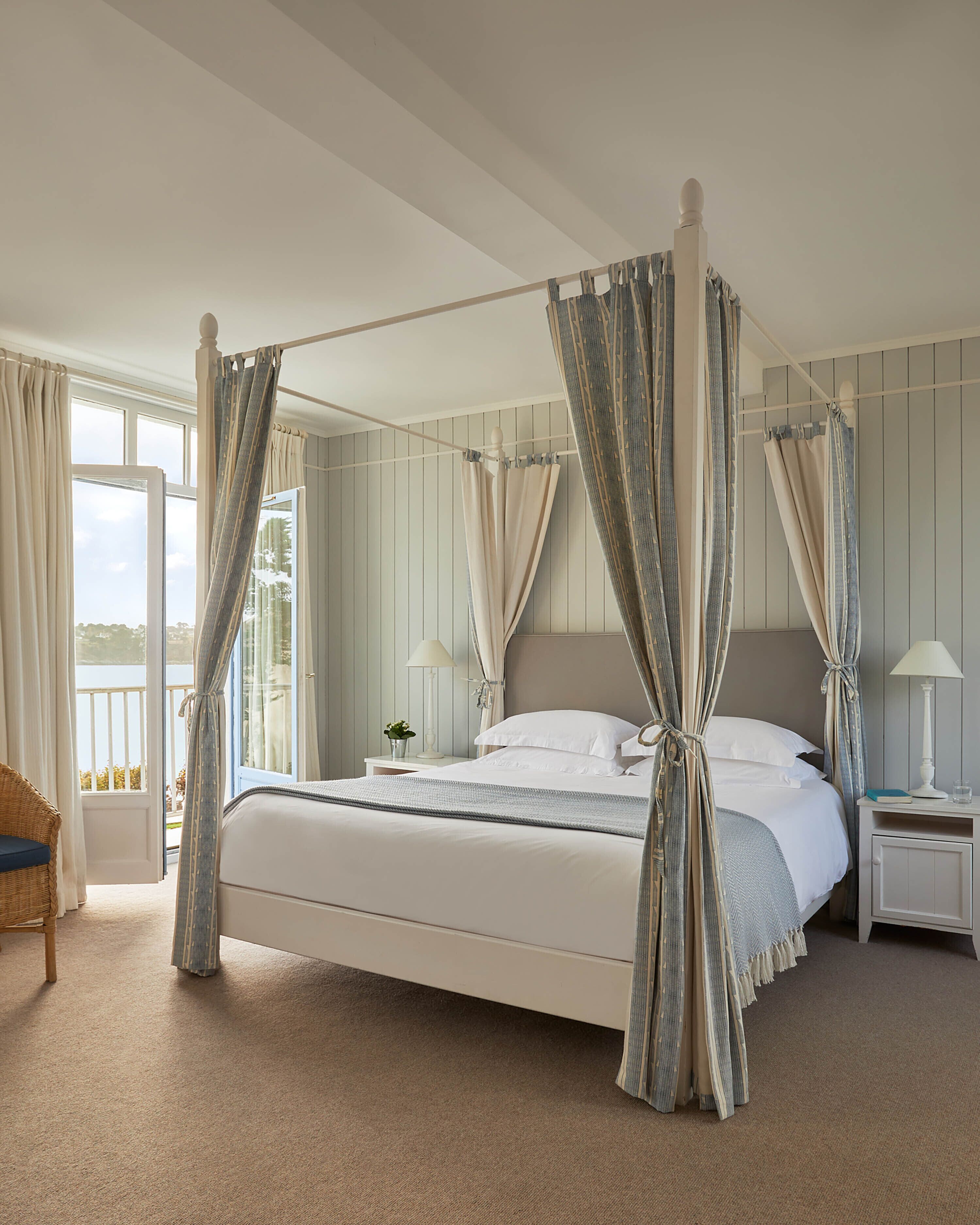 A four-poster bed with curtains, a nightstand with a lamp, and a view of the outdoors.