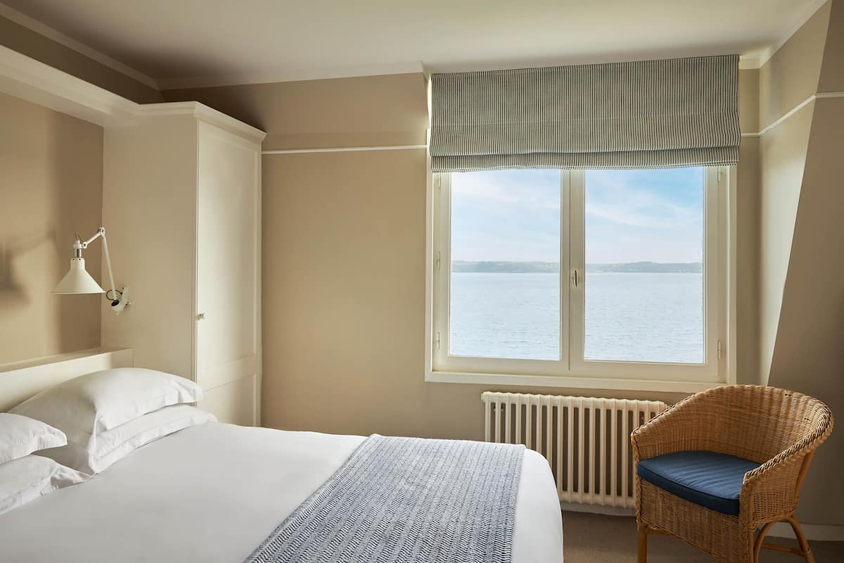 A bedroom with a large bed, a wicker chair, a window with a view of the sea, and a radiator.