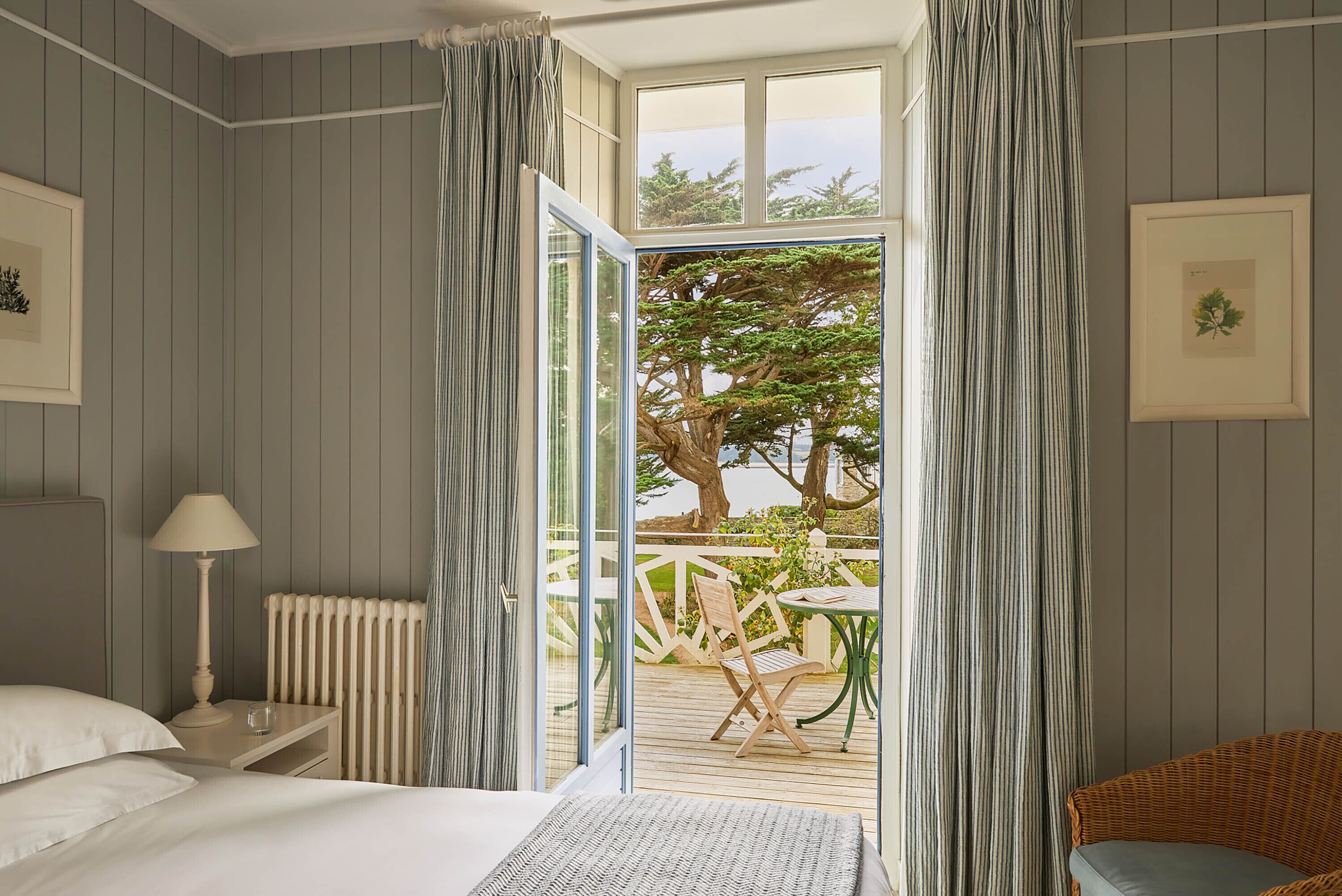 A bedroom with a bed, bedside table, lamp, and a view of an outdoor deck with a chair and table.