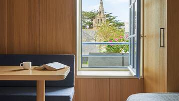 Una habitación con un banco de madera, una mesita con una taza y un libro, una cama con una manta azul y una ventana con vistas a una iglesia y follaje.