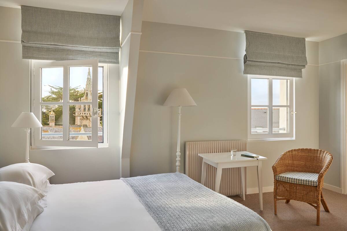A bedroom with a bed, a chair, a table, and two windows with curtains.