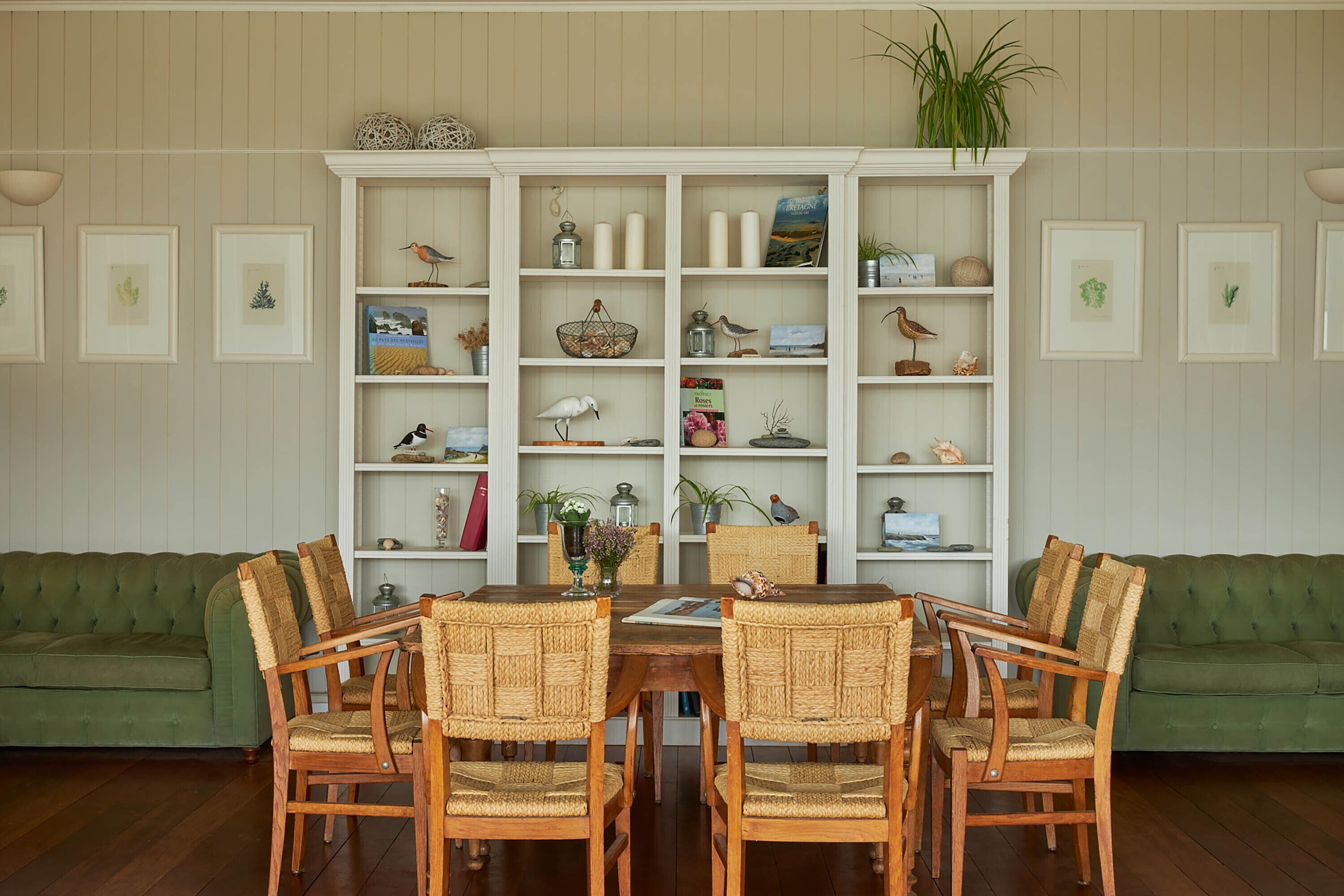 A dining area with a round wooden table and wicker chairs, flanked by green sofas. A white shelving unit with various decorative items and framed pictures is behind the table.