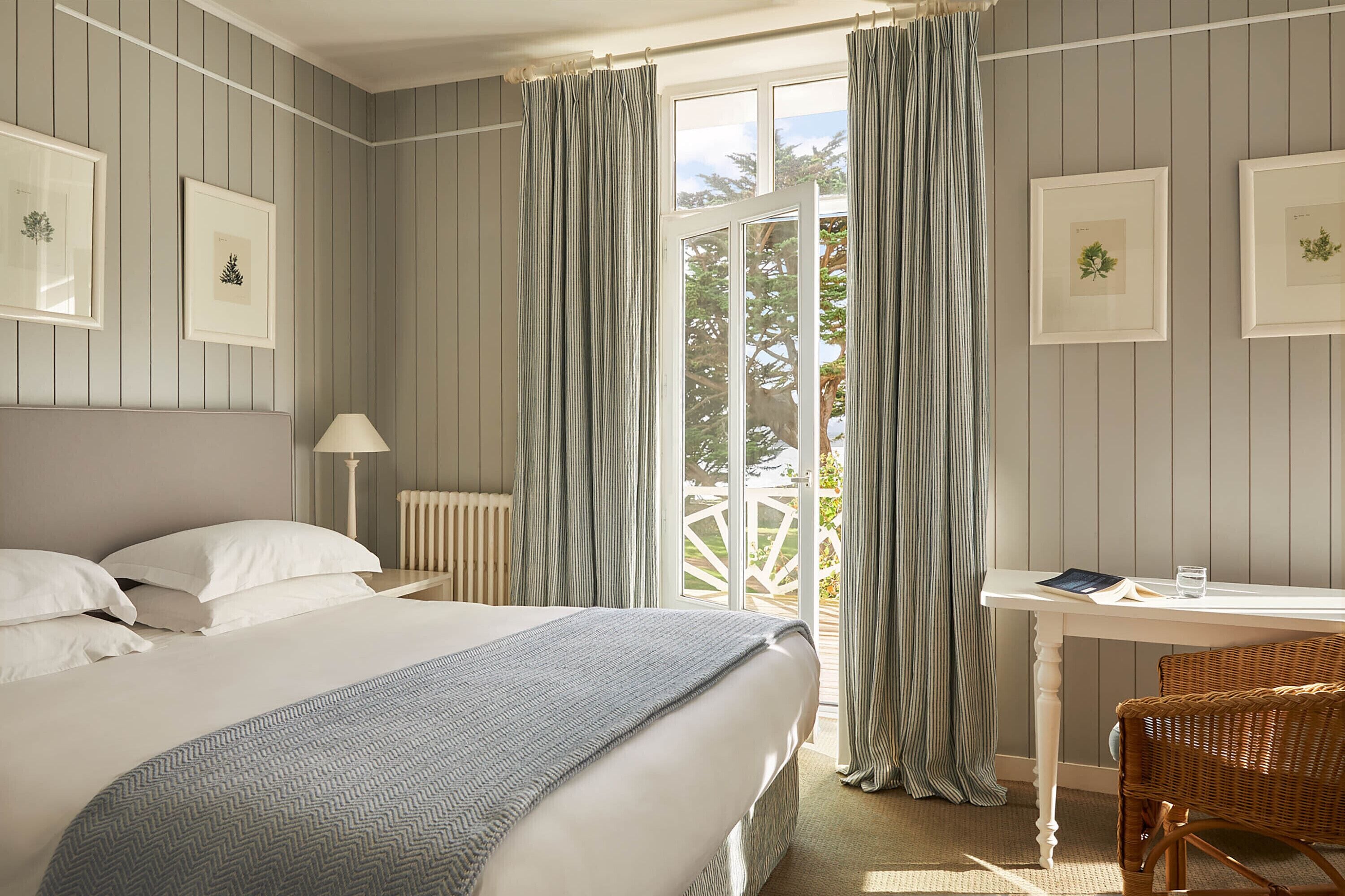 A bedroom with a bed, a bedside table with a lamp, a desk with a chair, and a view of the outdoors through a door.