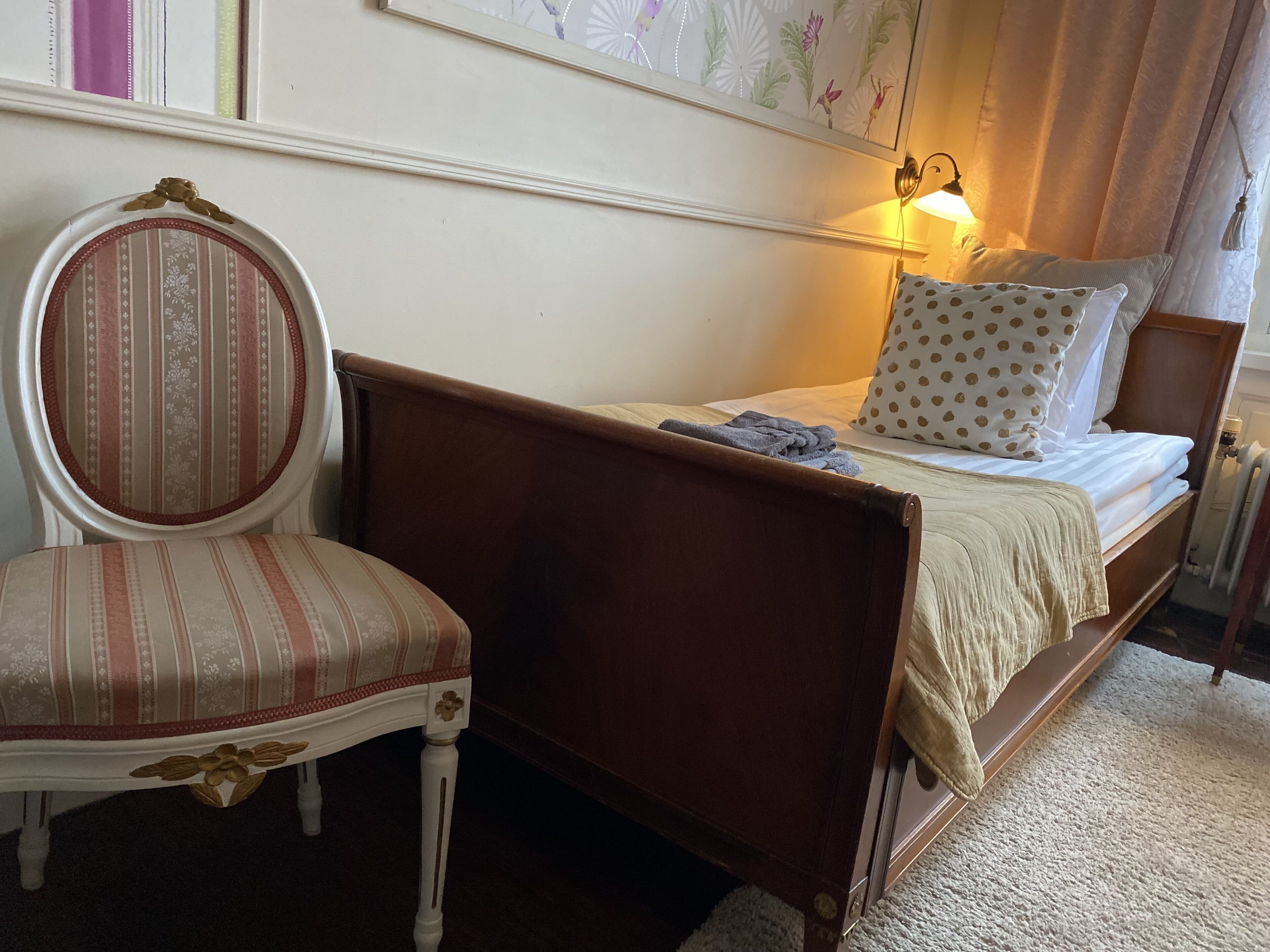 Una camera da letto con un letto di legno, una sedia a righe, una lampada a parete e un quadro floreale.