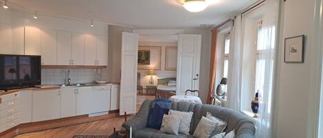 A modern living room with a grey sofa, a coffee table, and a TV. A kitchen with white cabinets and a dining area with a table and chairs are visible in the background.