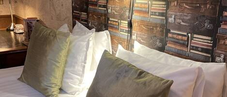 Una cama bien hecha con sábanas blancas y almohadas verdes, una mesita de noche con lámpara y una pared decorada con maletas antiguas.