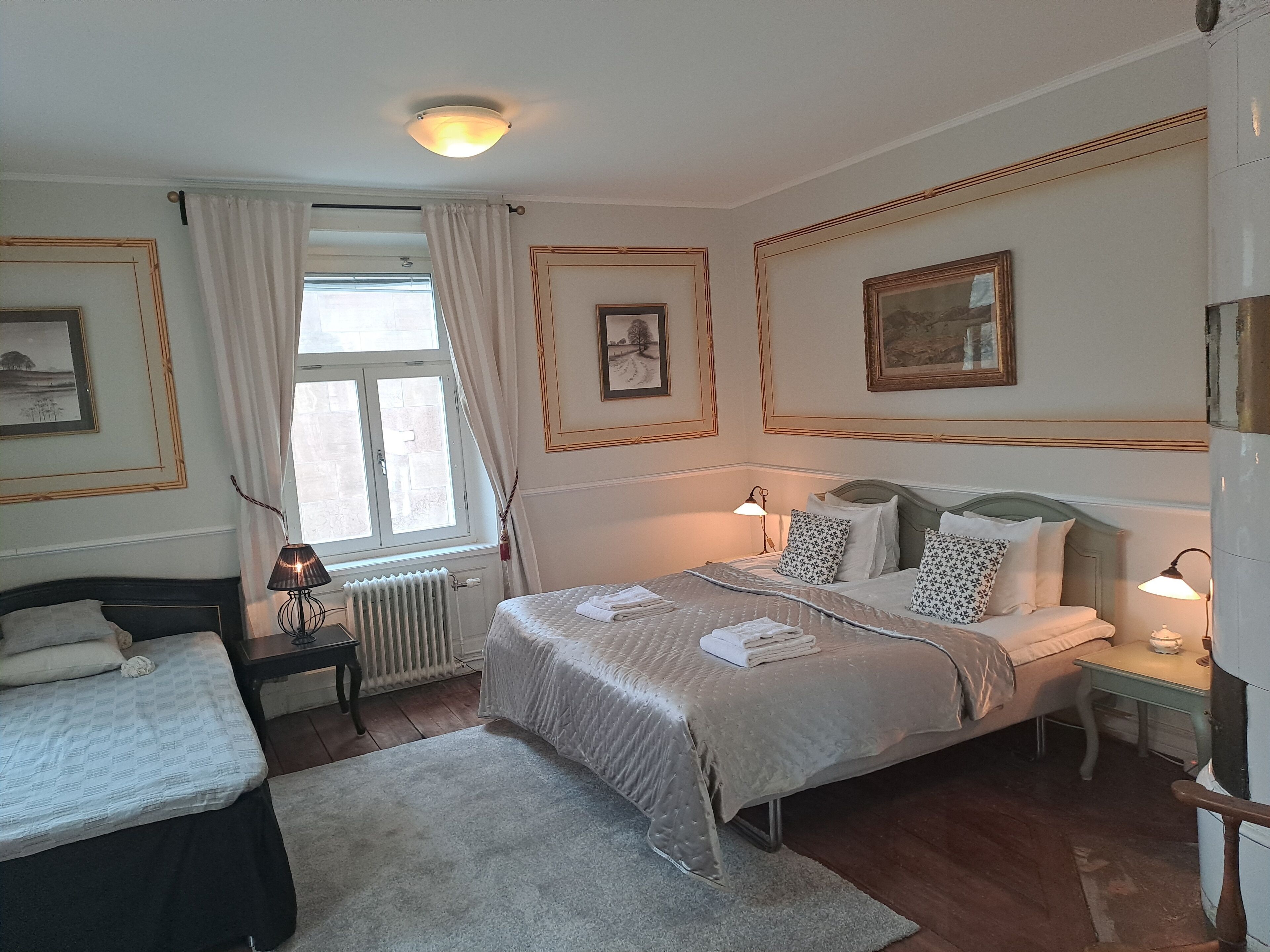Una habitación de hotel con una cama, una cama individual, un radiador, una ventana con cortinas y cuadros enmarcados en las paredes.