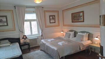 Una habitación de hotel con una cama, una cama individual, un radiador, una ventana con cortinas y cuadros enmarcados en las paredes.
