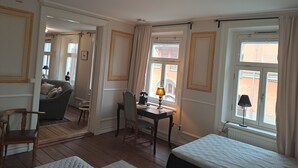 A hotel room with a bed, a desk with a lamp, a chair, a sofa, and large windows with curtains. - Strand Hotell (Norrköping)
