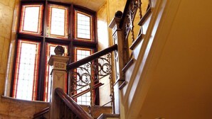 A grand staircase with ornate metal railings and a stone balustrade, leading to a landing with a large, arched window with stained glass. - Strand Hotell (Norrköping)