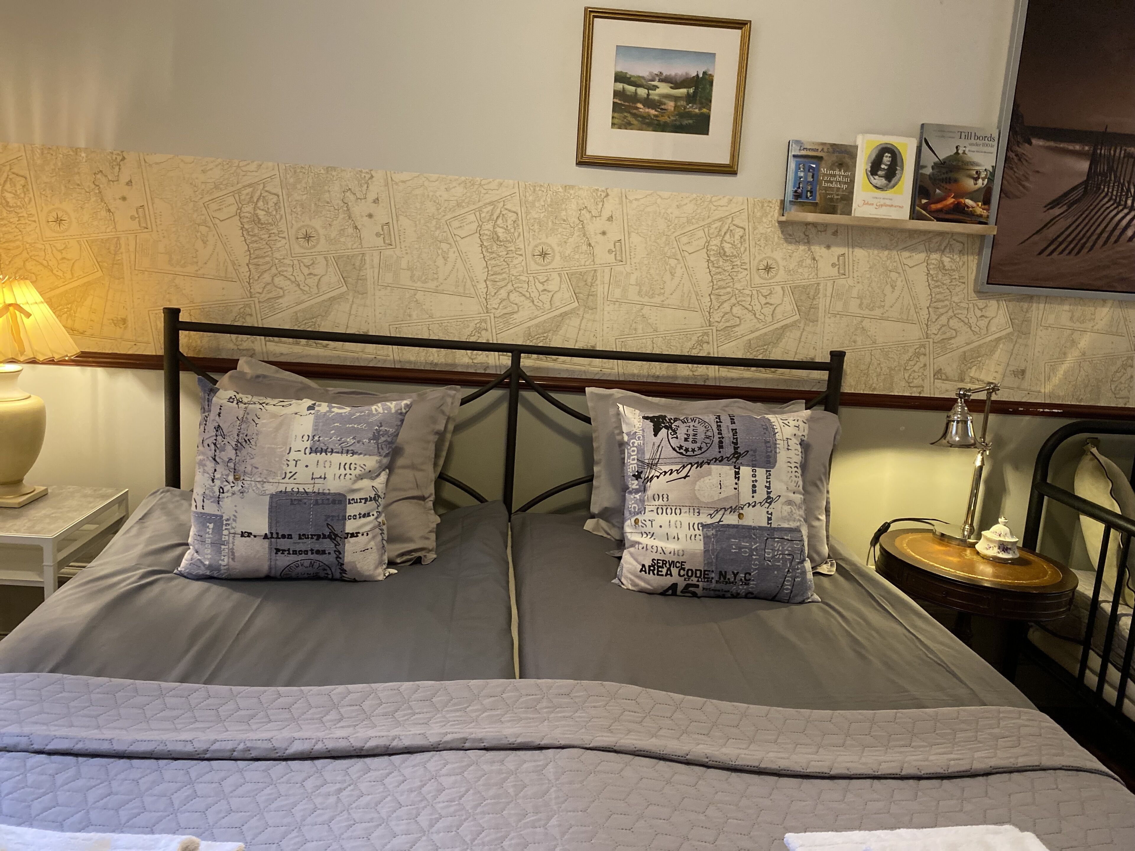 Una cama bien hecha con cubrecolcha acolchado, dos almohadas con diseños estampados, un cuadro enmarcado sobre la cama y una pequeña estantería con libros y una lámpara.