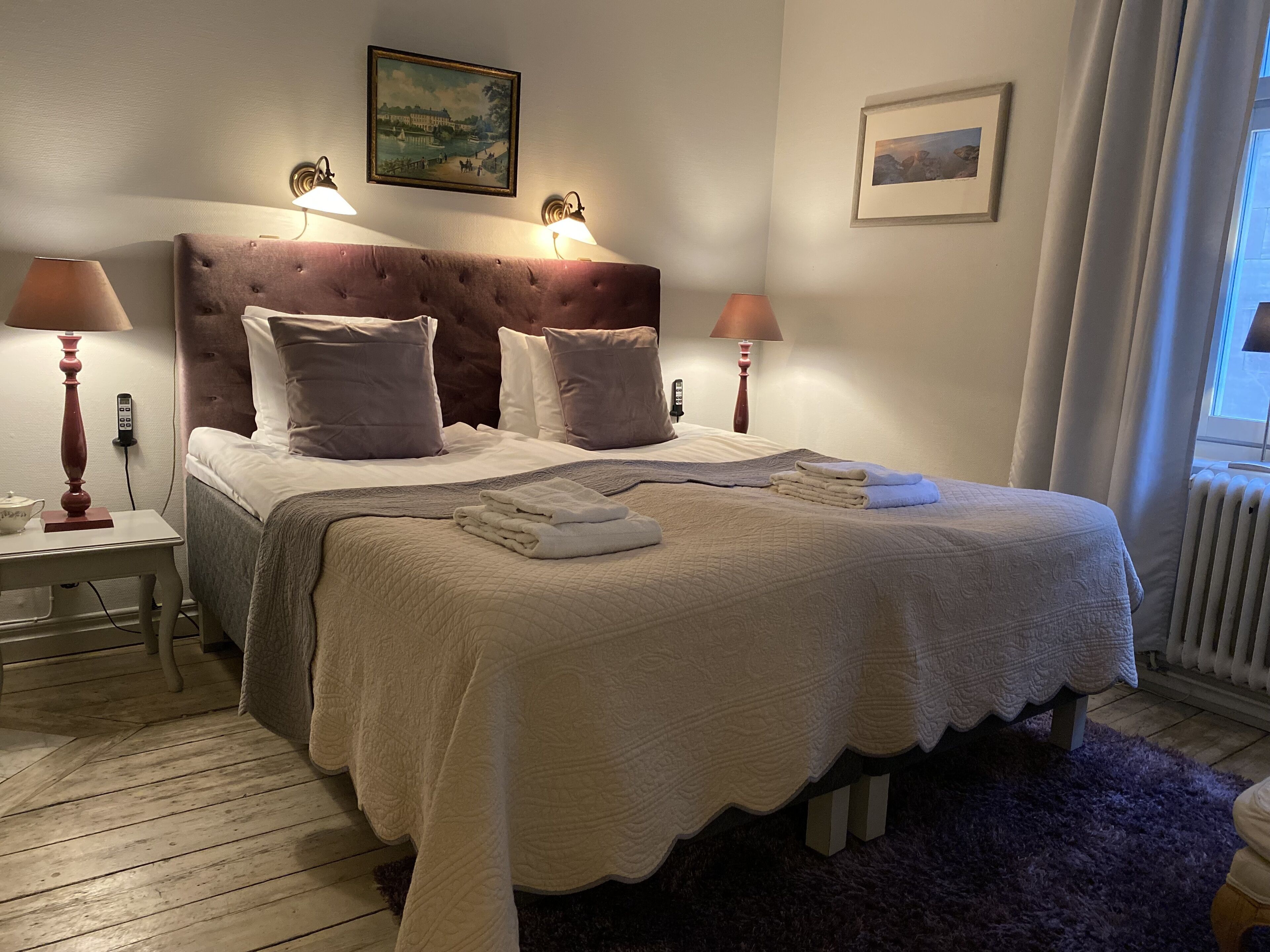 Una cama bien hecha con cubrecama blanco acolchado, dos lámparas de noche, un cuadro en la pared y una ventana con cortinas.