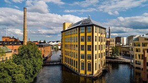 A yellow building with a corner turret, situated by a canal with industrial structures in the background. - Strand Hotell (Norrköping)