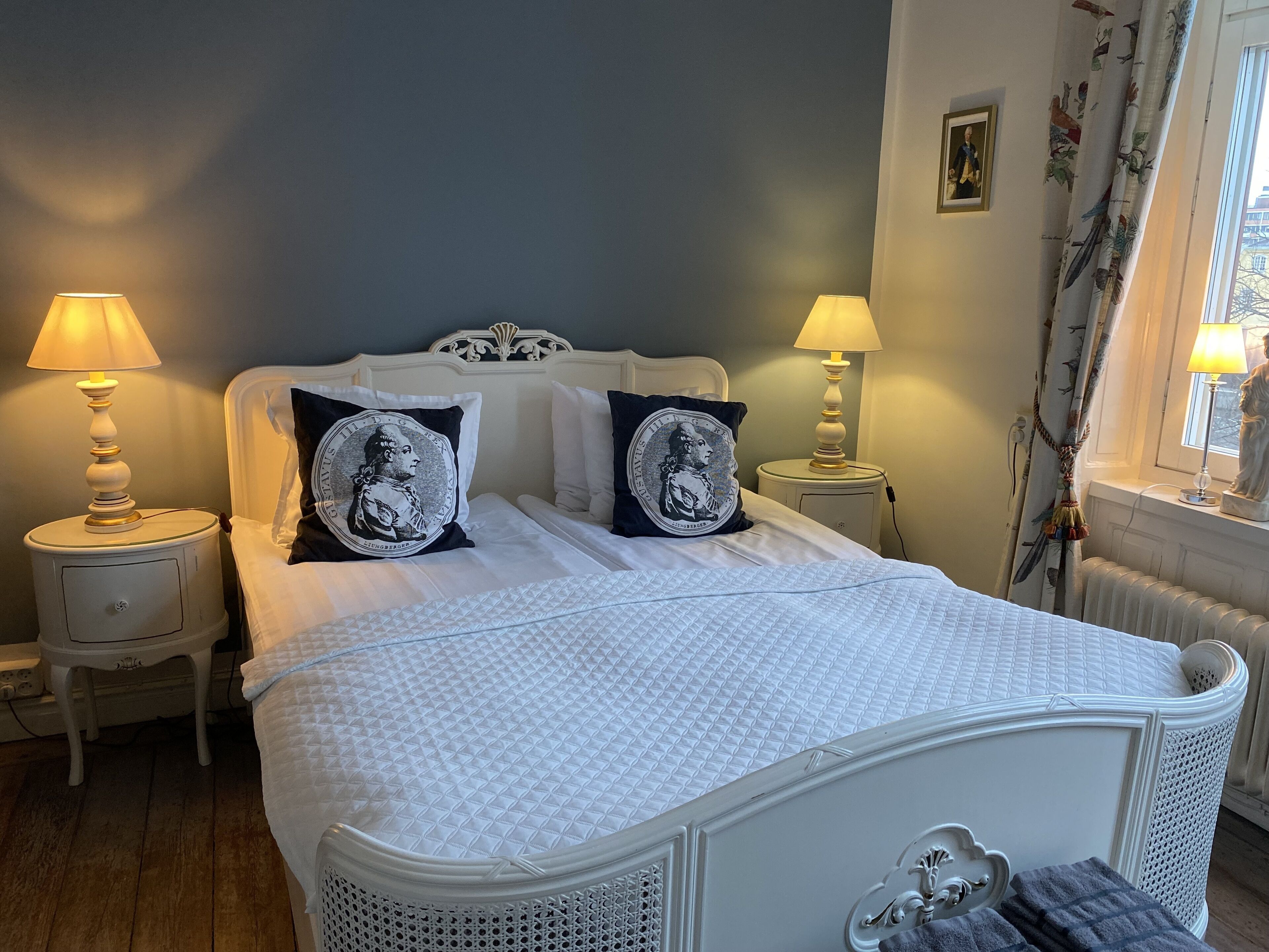Ein ordentlich bezogenes Bett mit weißer Bettwäsche und zwei Zierkissen, begleitet von zwei Nachttischen mit Lampen, in einem klassisch eingerichteten Zimmer.