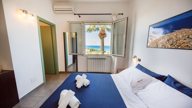 Ein Hotelzimmer mit Bett, Meerblick, einem Gemälde und einem Fenster mit Blick auf eine Palme.
