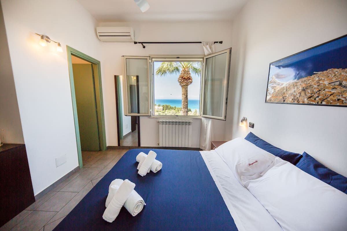 Une chambre d’hôtel avec un lit, une vue sur la mer, un tableau et une fenêtre donnant sur un palmier.