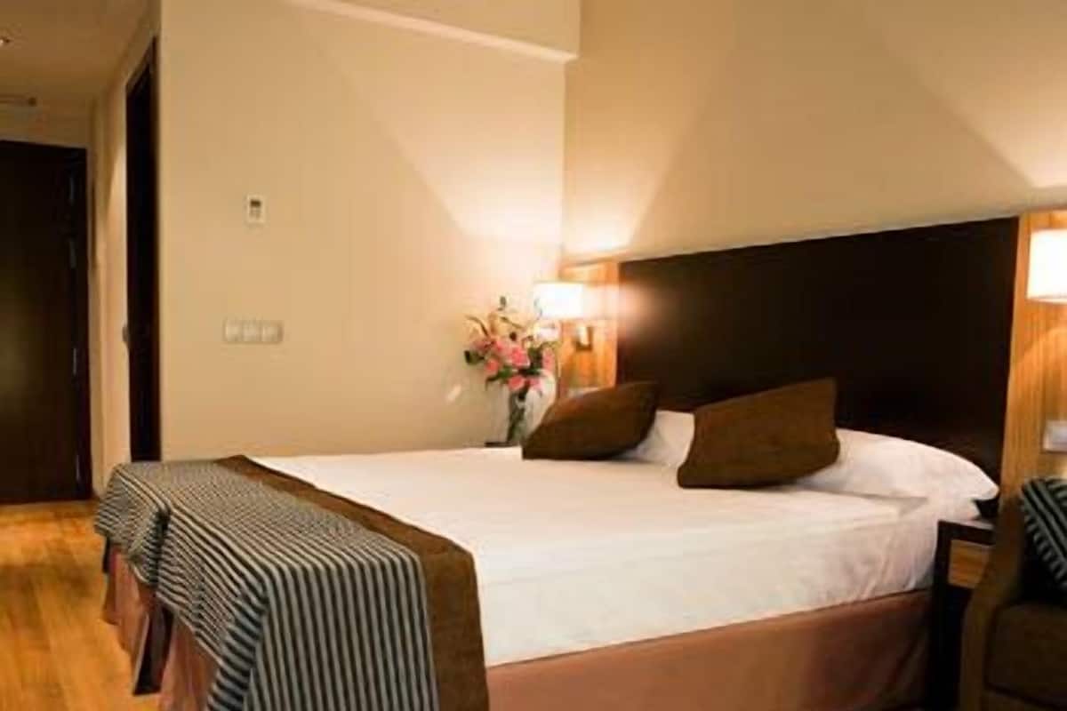 Ein Hotelzimmer mit einem großen Bett, Holzboden, einem Kopfteil, Nachttischlampen und einem kleinen Tisch mit Blumen in einer Vase.