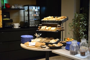 Disposição de buffet com diversos pães, doces e frutas.