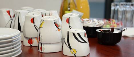 A table with white ceramic cups with abstract designs, plates, and a black bowl with utensils.