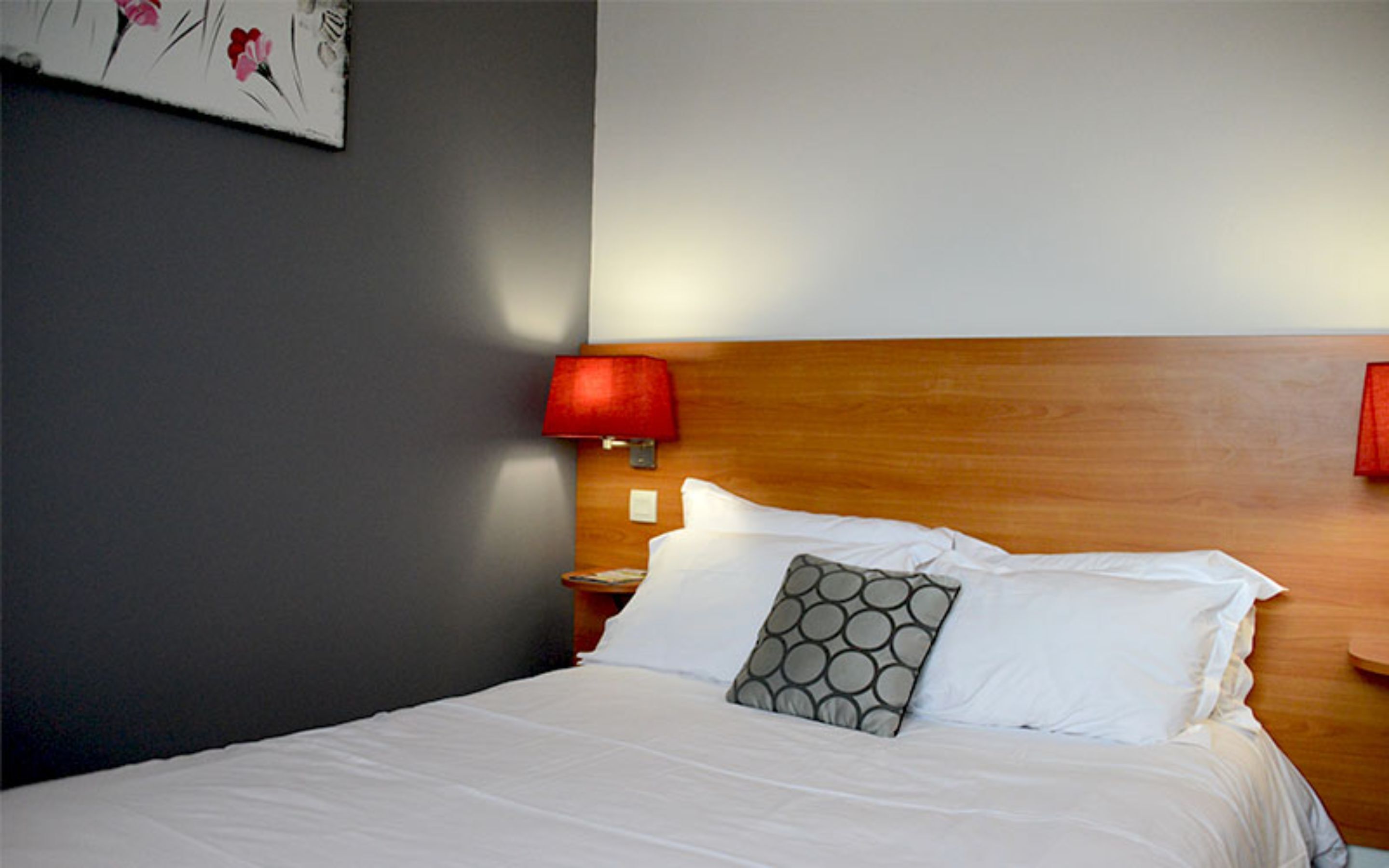 Een netjes opgemaakt bed met witte lakens, een houten hoofdbord, een rode bedlamp en een kunstwerk aan de muur.
