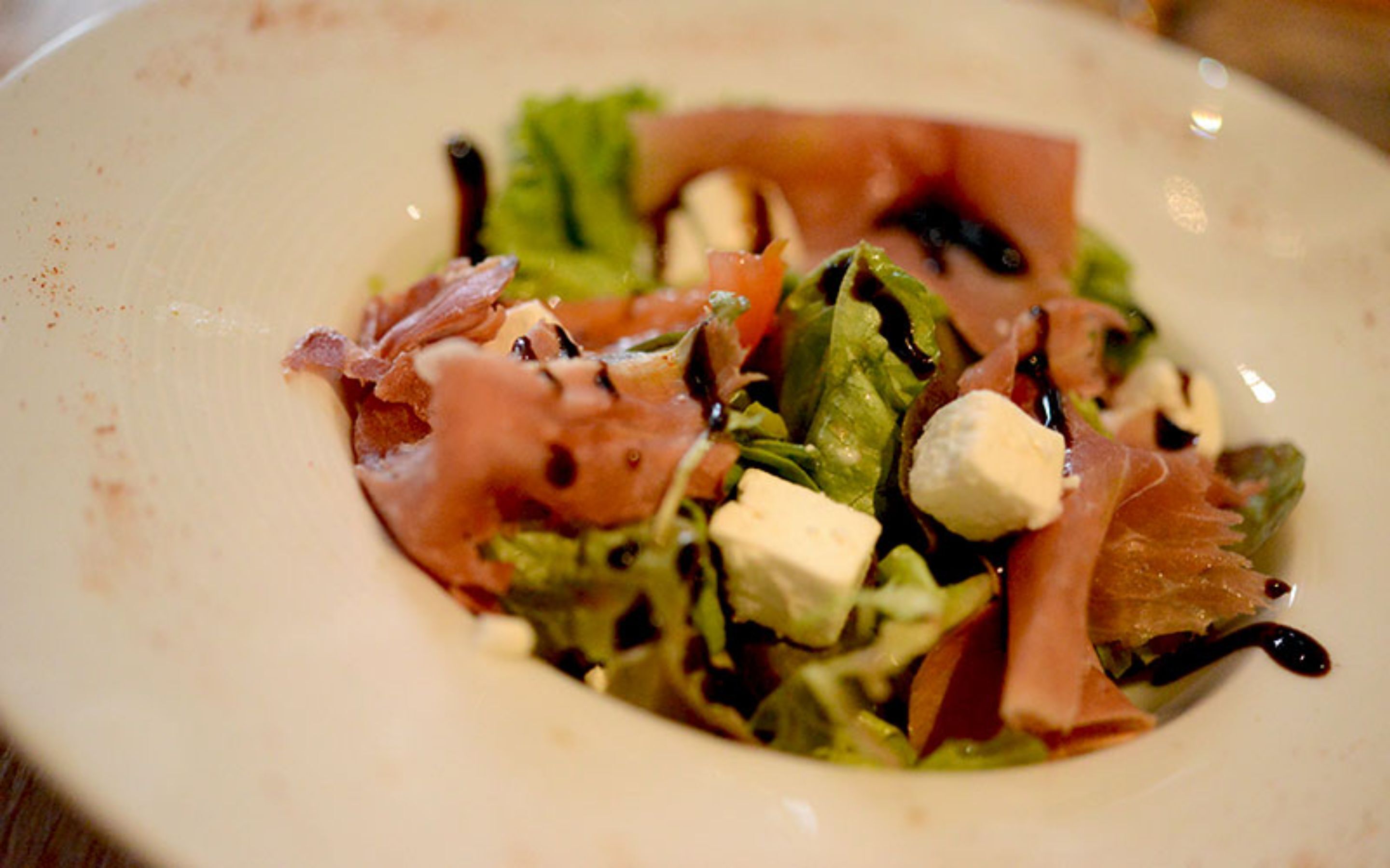 A plate with a salad consisting of greens, tomatoes, cheese, and prosciutto.