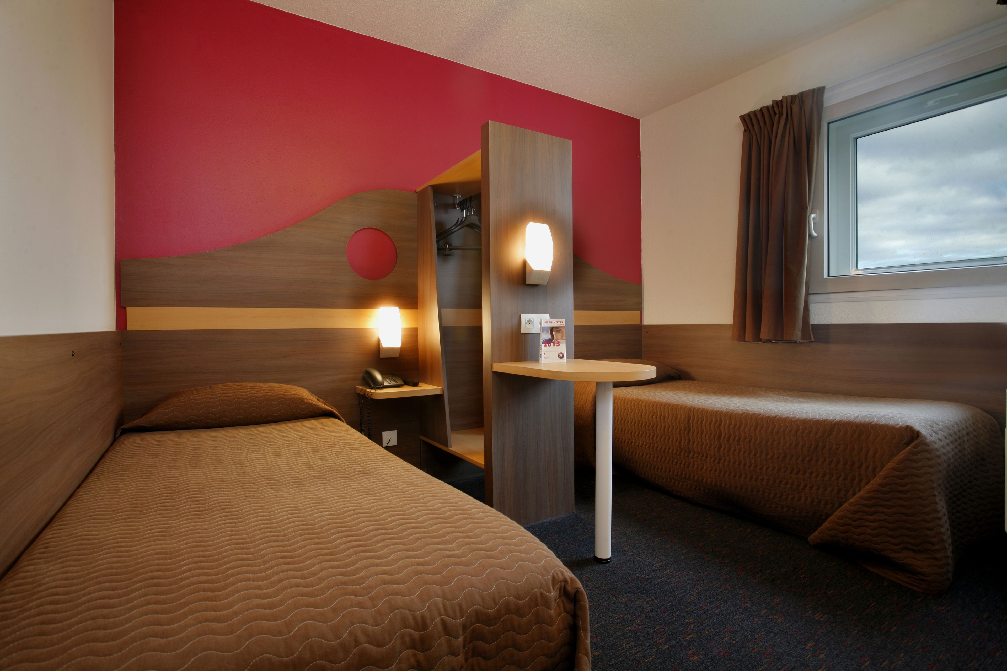 A hotel room with two beds, a small table, a mirror, and a window with curtains.