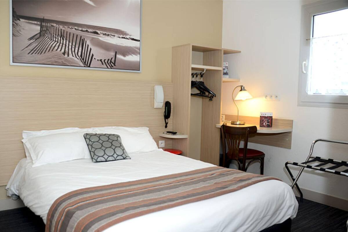 Una habitación de hotel con una cama, un escritorio con lámpara, una silla, un perchero y un teléfono.