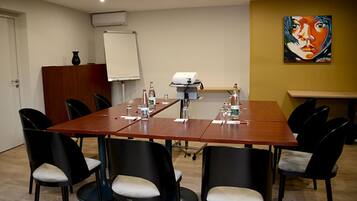 Una sala de conferencias con una larga mesa, sillas negras con cojines blancos, una pizarra, un equipo de aire acondicionado y un cuadro en la pared.