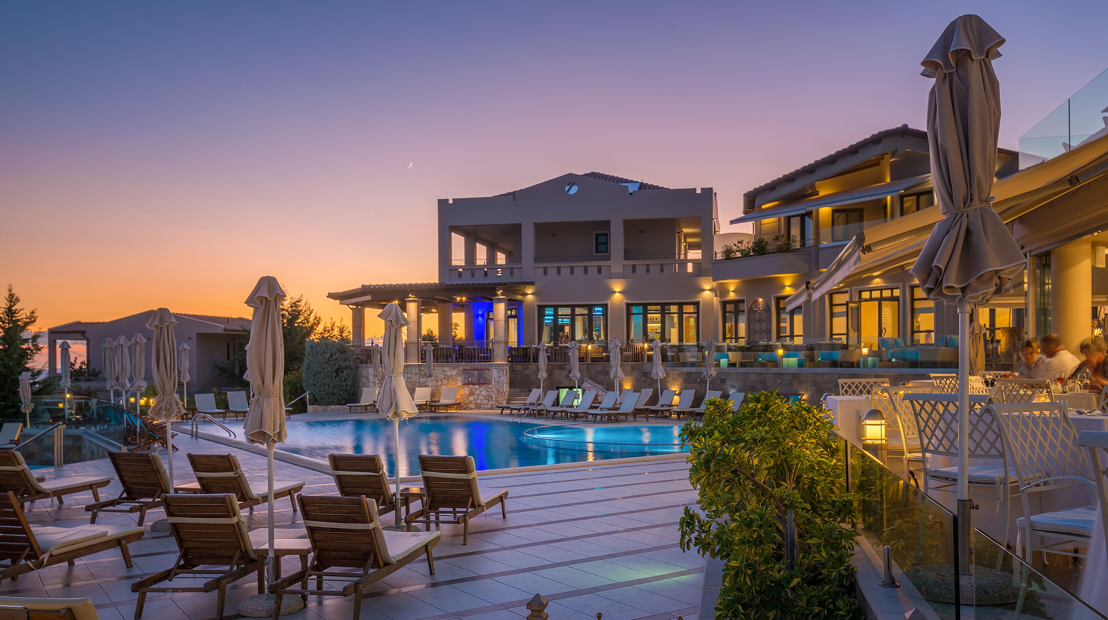 A poolside area with lounge chairs and umbrellas, a pool, and a building with multiple floors and outdoor seating.