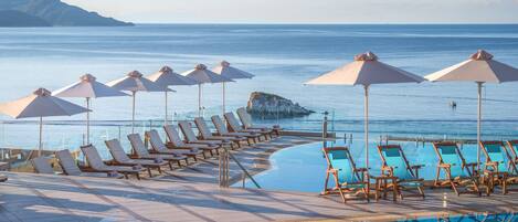 Zona de piscina con sillas de descanso y sombrillas, con vista a un mar tranquilo y un promontorio rocoso.