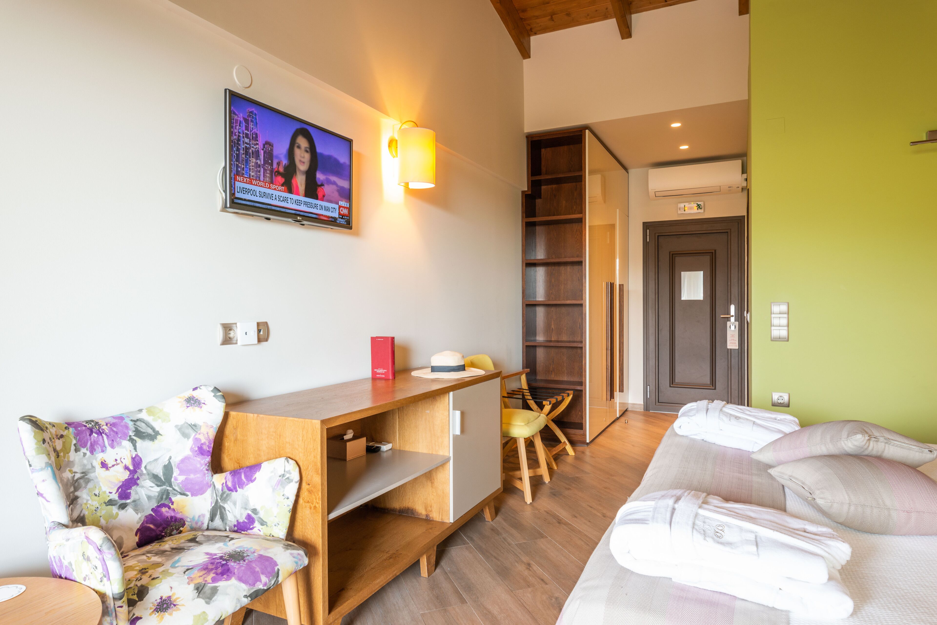 Une chambre d’hôtel avec un bureau en bois, un fauteuil à motifs floraux, un lit avec des oreillers, un téléviseur fixé au mur et un escalier menant à une autre pièce.