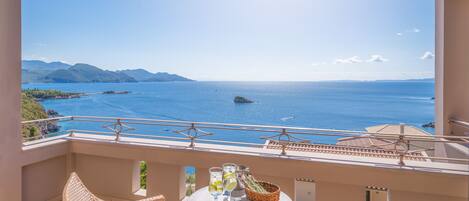 A balcony with wicker chairs, a small table with drinks, and a view of the sea and mountains.