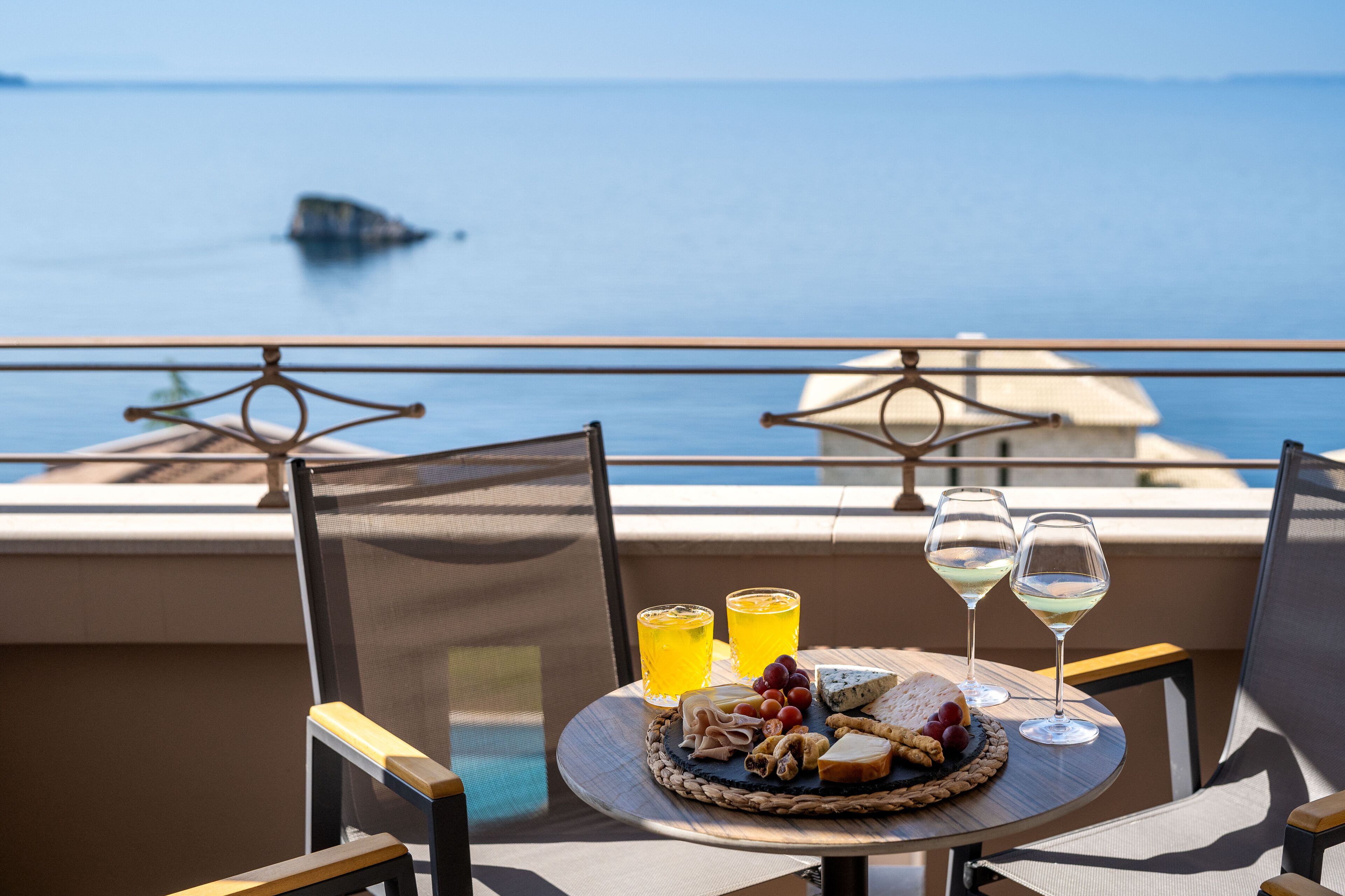 A table with a cheese platter, glasses of juice, and wine, set on a balcony overlooking a body of water.