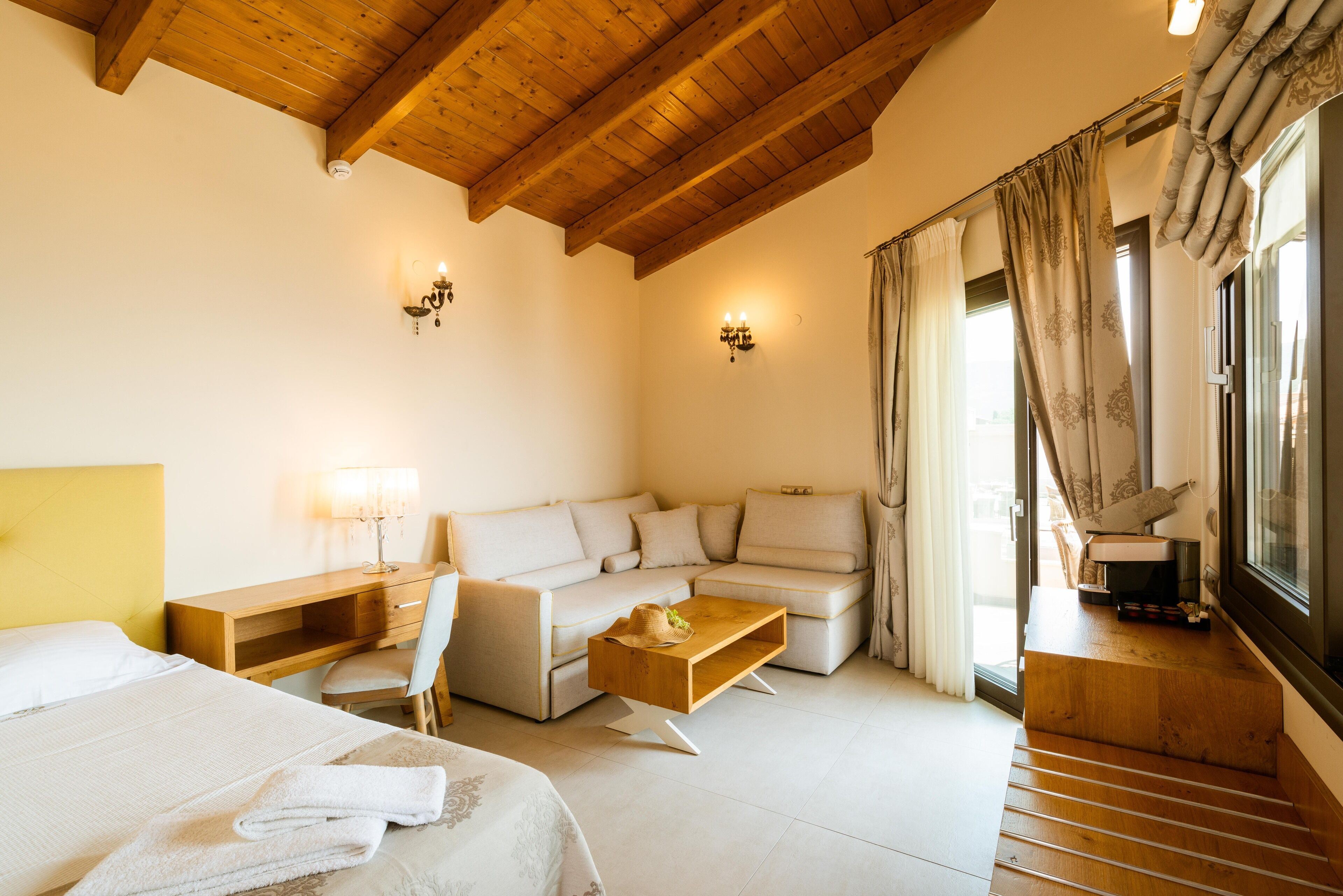 A modern hotel room with a wooden ceiling, a sofa, a bed, a desk, and a window with curtains.