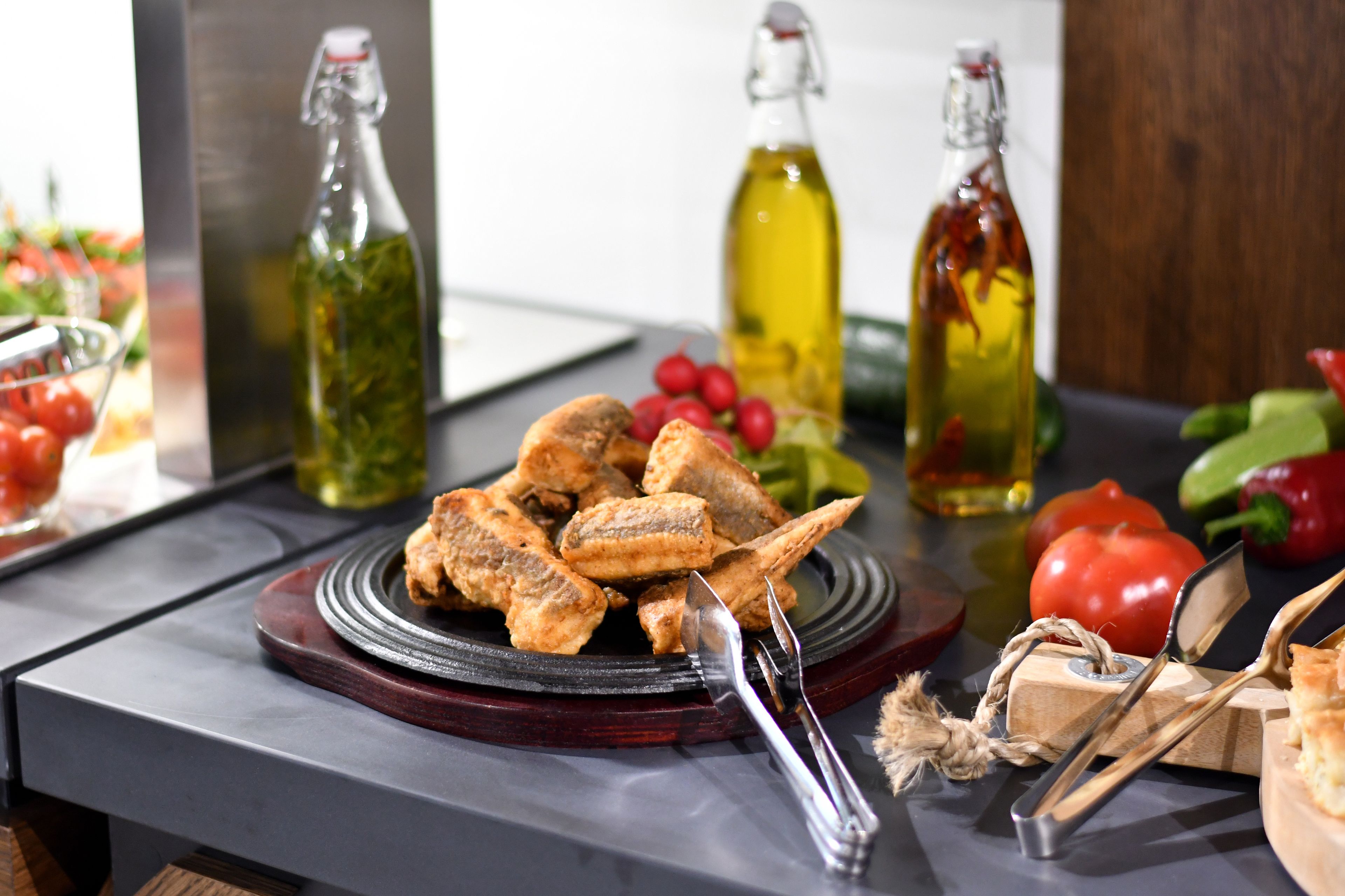 A dining table with a plate of fried fish, a bottle of olive oil, and various vegetables.