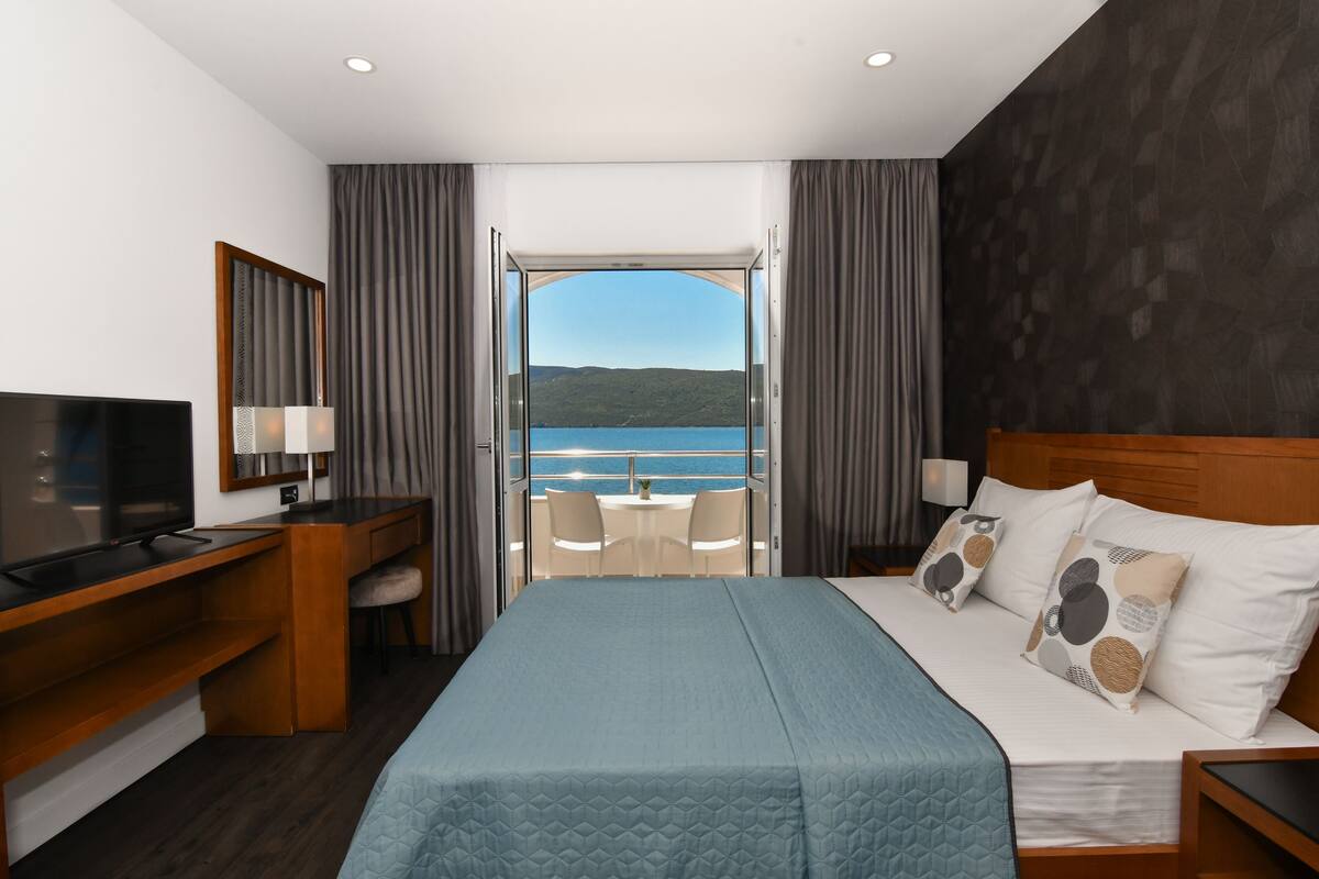A hotel room with a bed, a TV, a desk, and a balcony with a view of the sea.