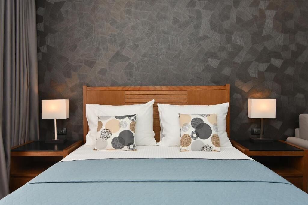 Ein ordentlich bezogenes Bett mit blauer Bettdecke, zwei Kissen mit geometrischem Muster und je eine Nachttischlampe auf Holz-Nachttischen.