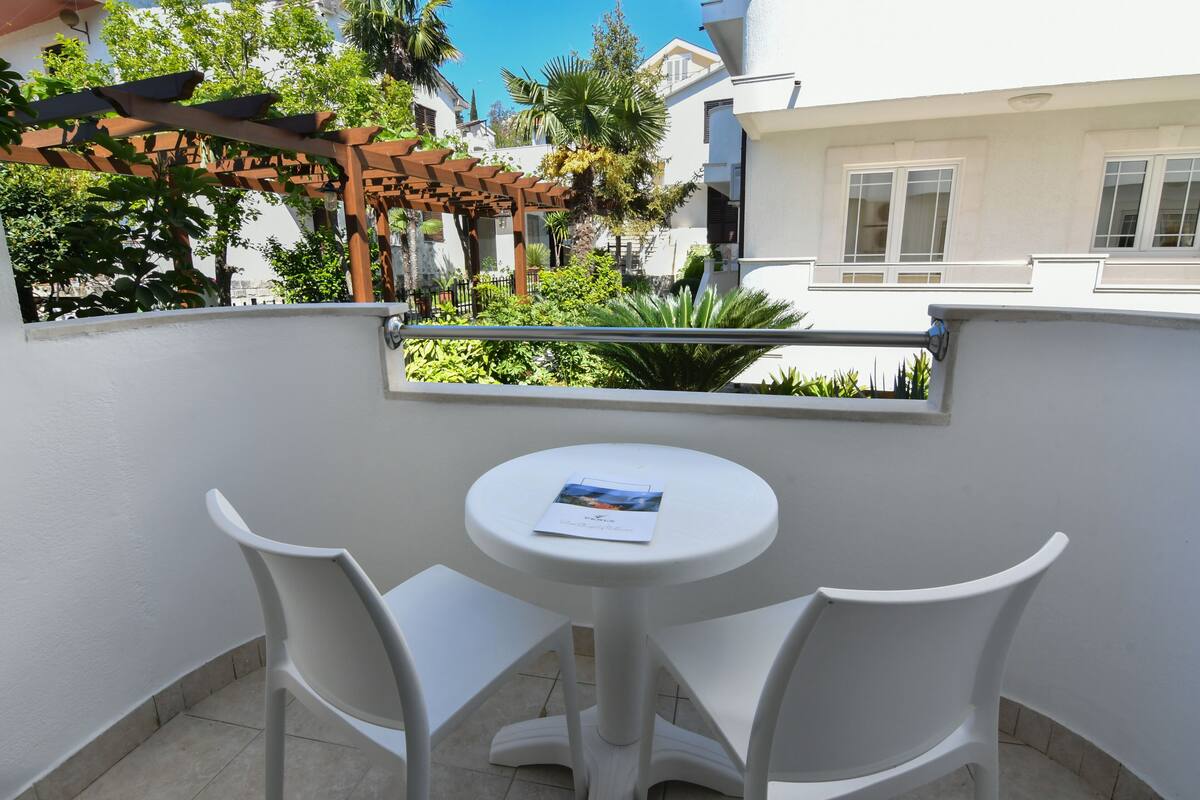A small outdoor patio with a white table and two white chairs, surrounded by a white wall and a wooden pergola.