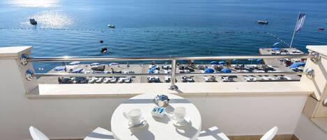 A balcony with a table set for two, overlooking a beach with boats and umbrellas.