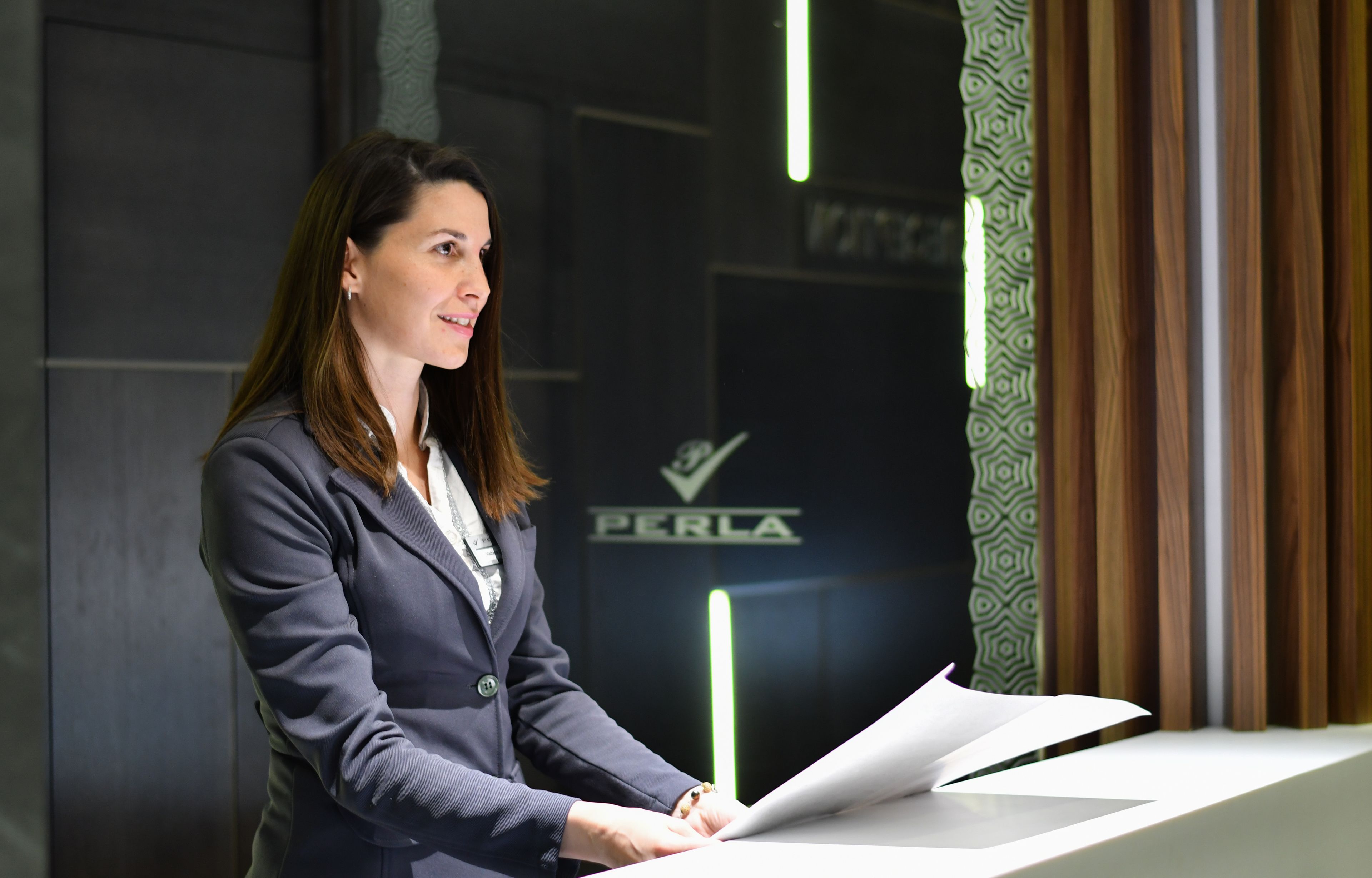 Een persoon in een pak zit aan een bureau met een document, in een kamer met een donkere muur en een logo met de tekst 'PERLA'.