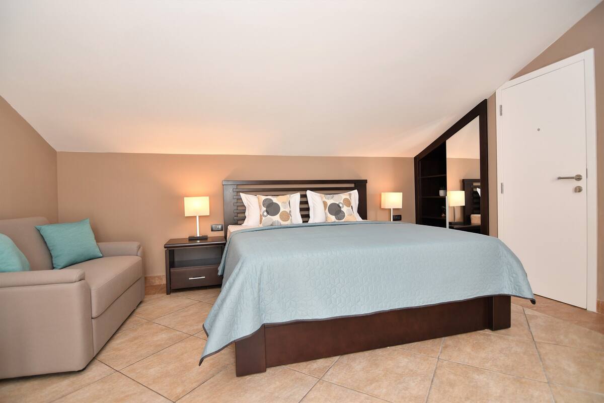 A bedroom with a bed, a nightstand, a lamp, a sofa, and a wardrobe.