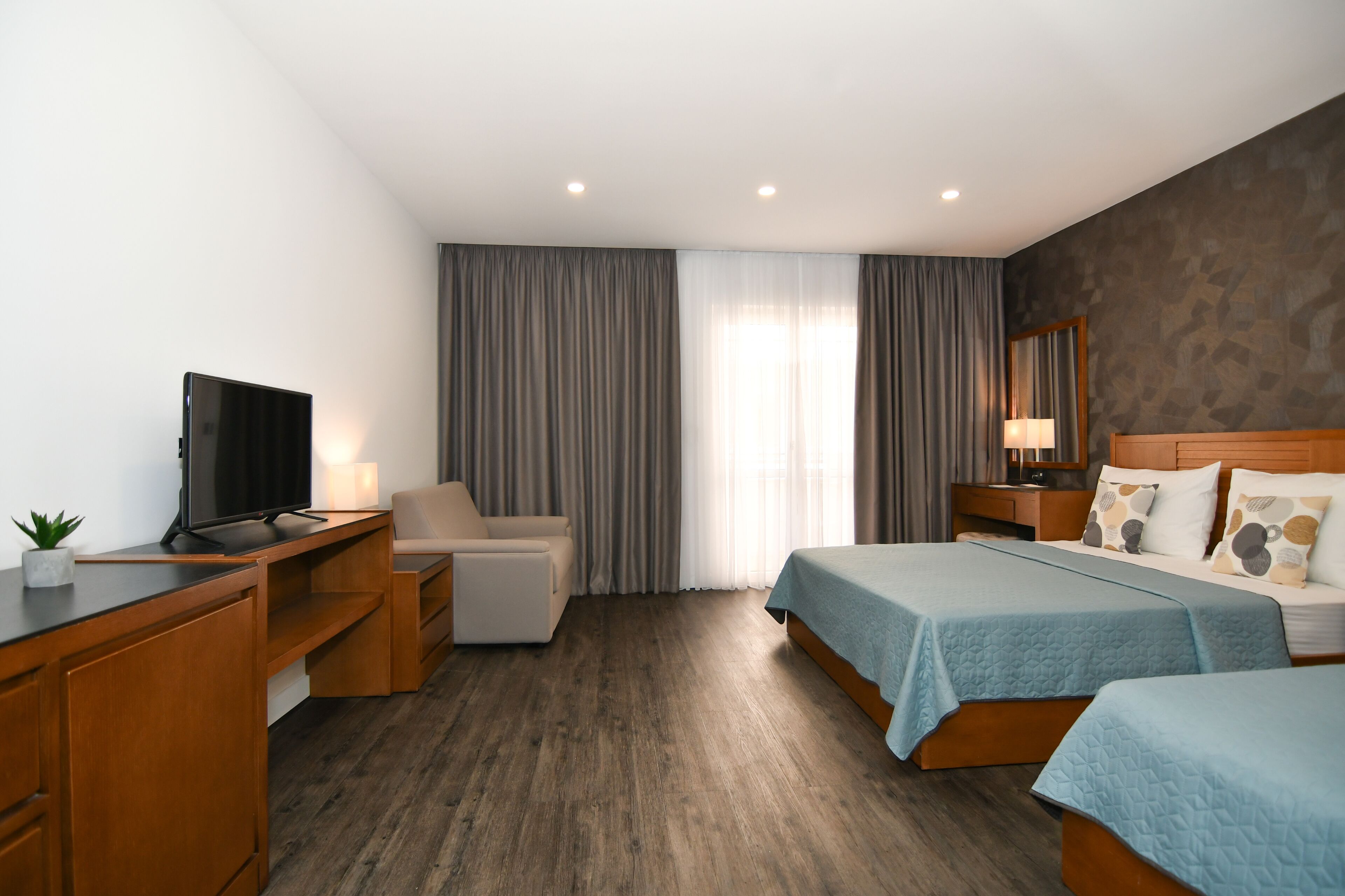 A hotel room with a wooden desk, a television, a sofa, two beds with blue bedding, a nightstand with a lamp, and a large window with curtains.