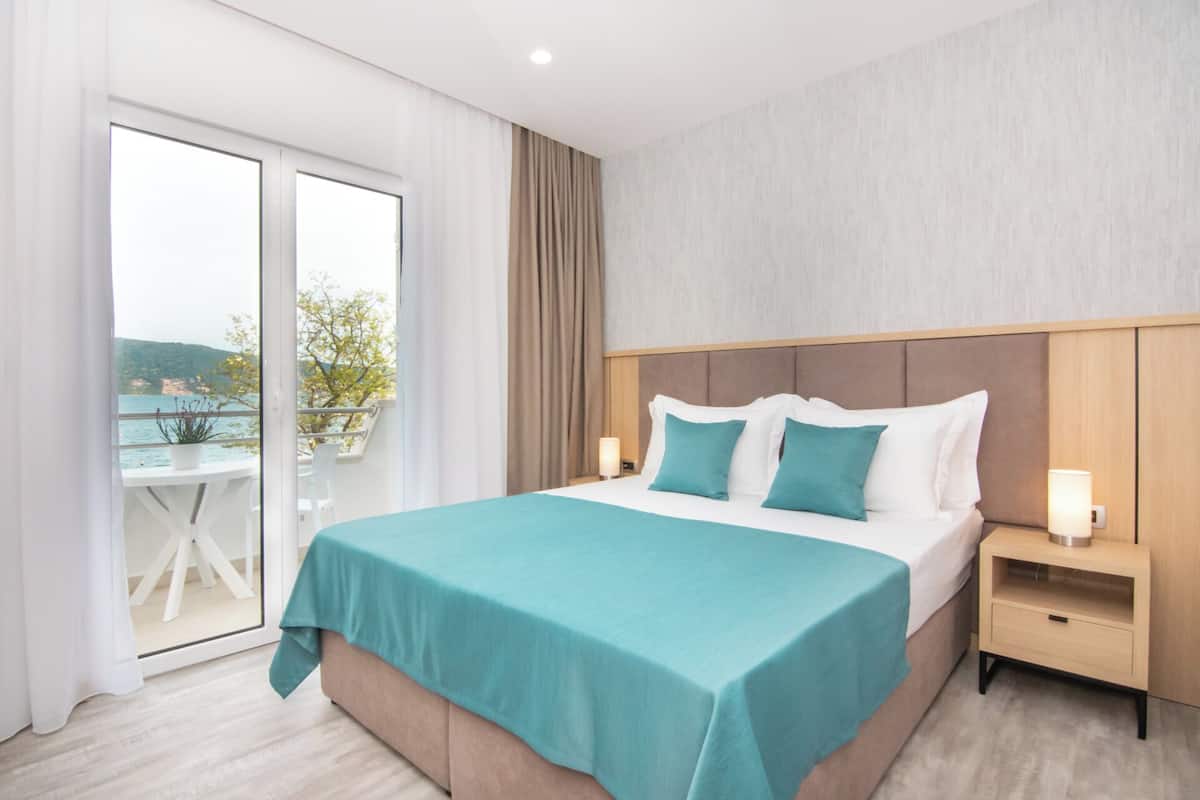 A hotel room with a bed, a nightstand, a table with a chair, and a view of the sea.