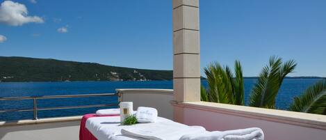 A balcony with a view of the sea, a massage table with towels, and a bottle of lotion.