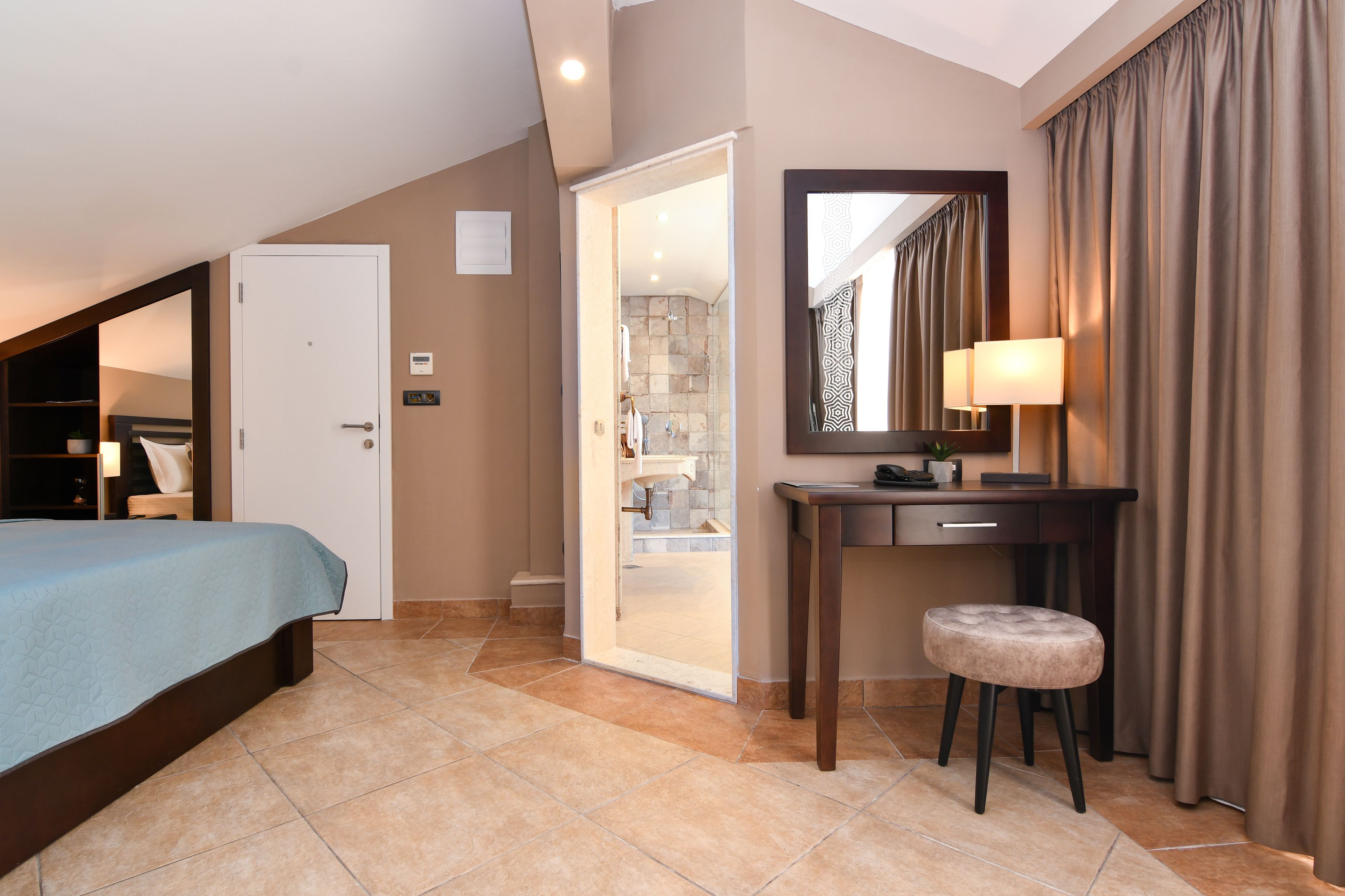 A bedroom with a bed, a desk, a chair, a mirror, and a bathroom visible through an open door.